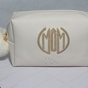 May include: White zippered cosmetic bag with a gold embroidered monogram "MOM" and a white pom-pom zipper pull.