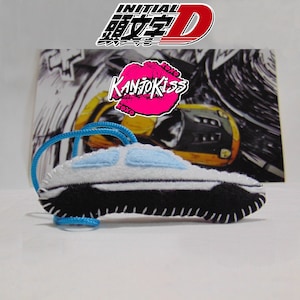 Op de afbeelding: Een witte en blauwe pluche autosleutelhangertje met zwarte details en een blauwe koord. De sleutelhangertje is voor een zwart-wit manga-stijl afbeelding met de tekst "Initial D" en "KanjoKiss" in roze.