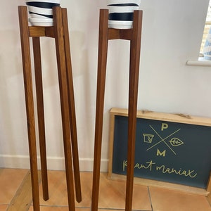 May include: Two wooden plant stands with black and white striped pots on top. The stands are made of dark brown wood and have a simple design. The stands are on a tiled floor. A sign with the text "Plant Maniac" is visible in the background.