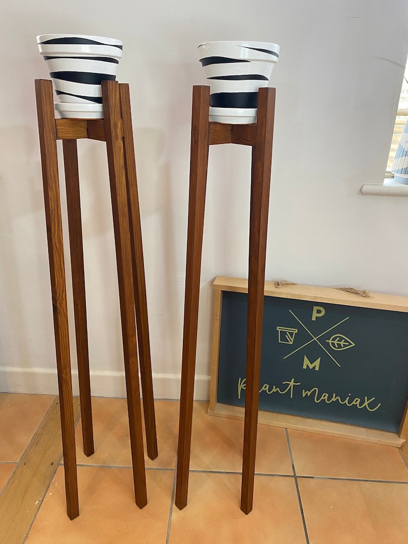 May include: Two wooden plant stands with black and white striped pots on top. The stands are made of dark brown wood and have a simple design. The stands are on a tiled floor. A sign with the text "Plant Maniac" is visible in the background.