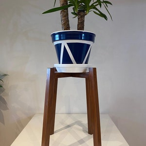 May include: A potted Yucca plant in a navy blue and white geometric designed planter, sitting on a wooden plant stand. The stand has a square top and four legs. The plant has multiple green leaves.