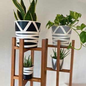 May include: A wooden plant stand with four shelves, holding three potted plants. The plants are a snake plant in a black and white geometric pot, a pothos in a gray and white geometric pot, and a succulent in a black and white striped pot.