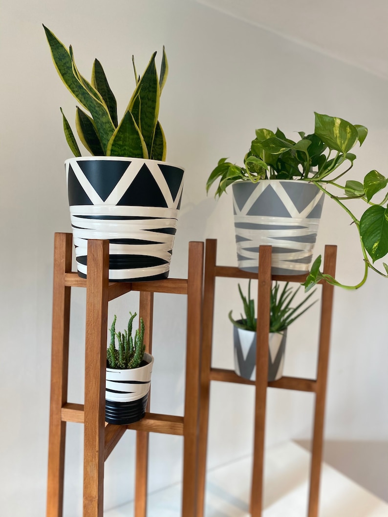 May include: A wooden plant stand with four shelves, holding three potted plants. The plants are a snake plant in a black and white geometric pot, a pothos in a gray and white geometric pot, and a succulent in a black and white striped pot.