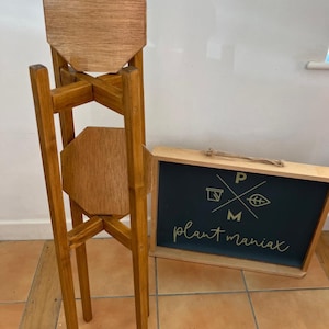 May include: A wooden plant stand with three octagonal shelves. The stand is stained a light brown color and has a rustic look. The stand is on a tiled floor. A sign with the text "plant maniac" is in the background.