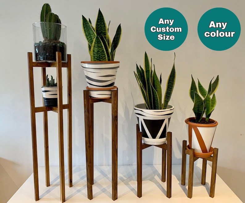 May include: Four wooden plant stands of varying heights, each displaying a potted plant. The plants include a cactus, a snake plant, and two further snake plants. The stands are all crafted from dark wood and feature a simple design. The text "Any Custom Size" and "Any colour" is visible in the image.