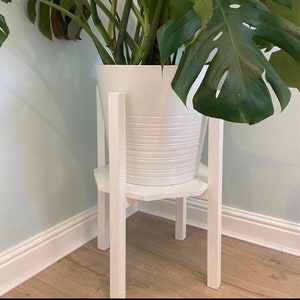 Pode incluir: Um suporte para plantas branco com quatro pernas e um topo hexagonal. Um vaso branco com uma planta verde de folhas está colocado no topo do suporte.