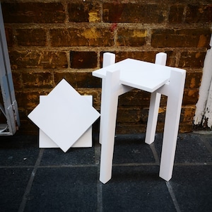 May include: White wooden stool with a square top and four legs. The stool is sitting on a black tiled floor.