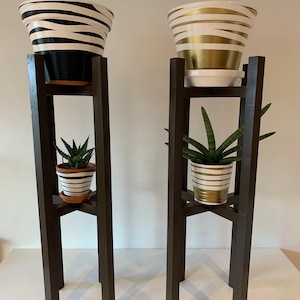 Könnte beinhalten: Zwei schwarze Pflanzenständer aus Holz mit zwei Topfpflanzen. Die oberen Blumentöpfe sind schwarz-weiß gestreift und gold-weiß gestreift. Die unteren Blumentöpfe sind weiß-schwarz gestreift und weiß-gold gestreift.