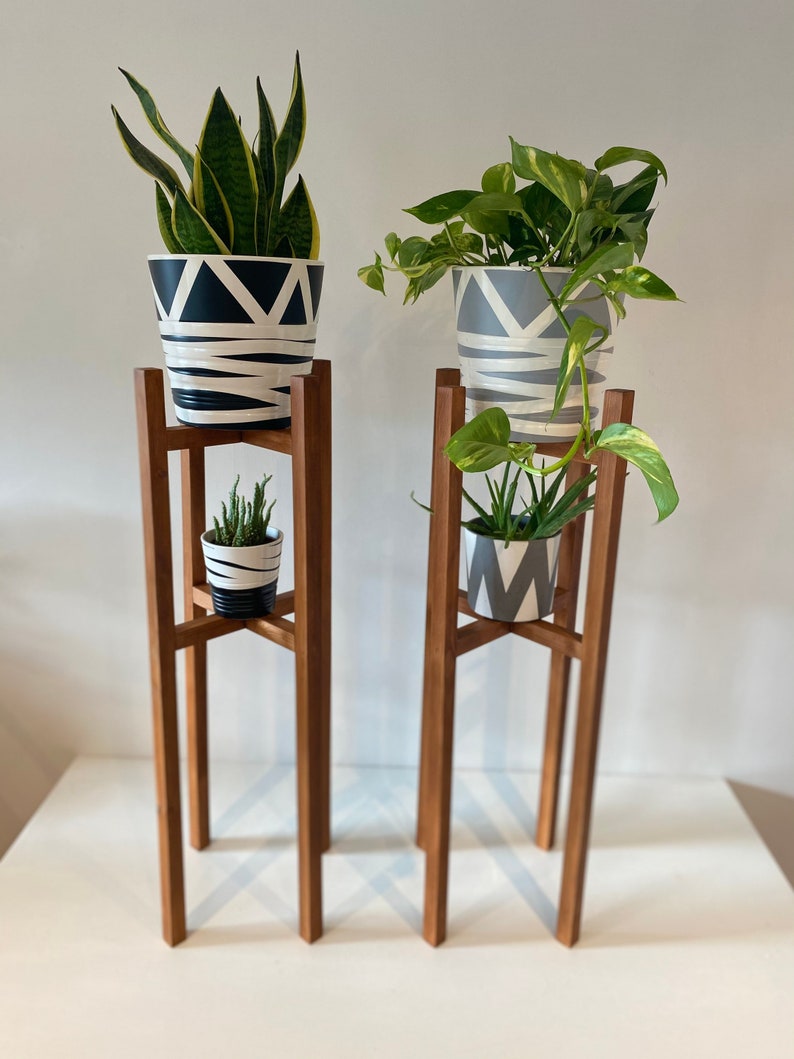 May include: Two wooden plant stands with three tiers each. The top tier of each stand holds a potted plant. The bottom tier of each stand is empty. The stands are made of natural wood and have a simple design.