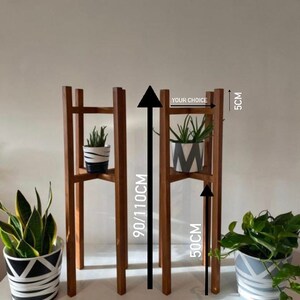 May include: Two wooden plant stands with two shelves. The stands are 50 cm wide and 90/110 cm tall. The stands are made of natural wood and have a rustic look. The stands are perfect for displaying plants or other decorative items. "Your Choice" is written on the stand.
