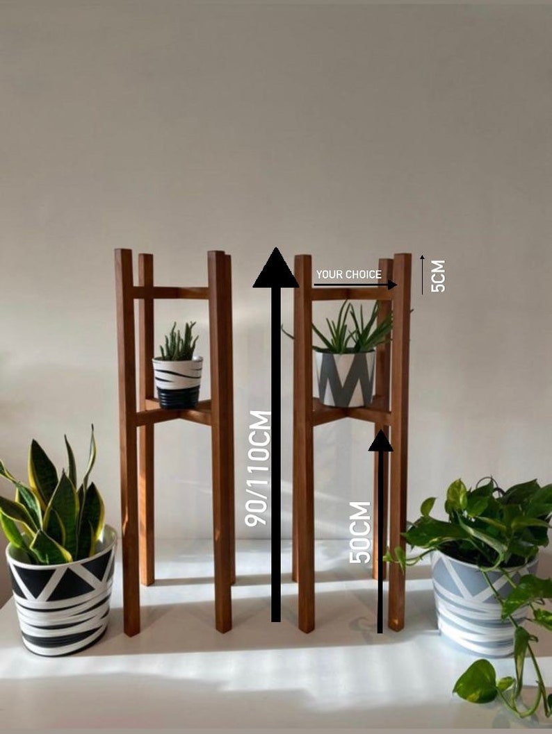 May include: Two wooden plant stands with two shelves. The stands are 50 cm wide and 90/110 cm tall. The stands are made of natural wood and have a rustic look. The stands are perfect for displaying plants or other decorative items. "Your Choice" is written on the stand.