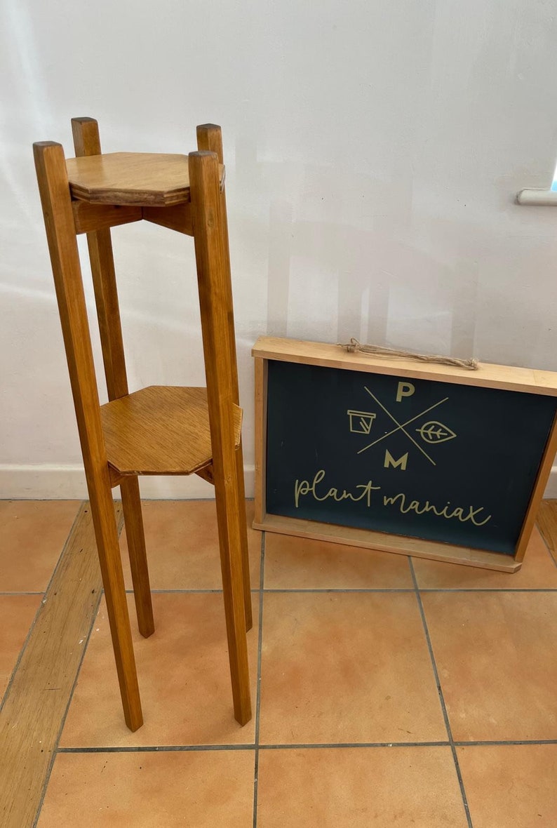May include: A two-tiered wooden plant stand with a hexagonal top and bottom. The stand is made of natural wood and has a rustic finish. The stand is tall and slender, with four legs.