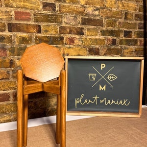 May include: A wooden plant stand with a hexagonal top. The stand is made of natural wood and has a rustic look. The stand is in front of a brick wall with a sign that says "plant maniac" in gold lettering.