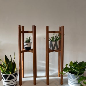 May include: Two wooden plant stands with three tiers each. The stands are made of dark brown wood and have a simple, modern design. Each stand has a potted plant on the top tier. The plants are in white pots with black geometric patterns.