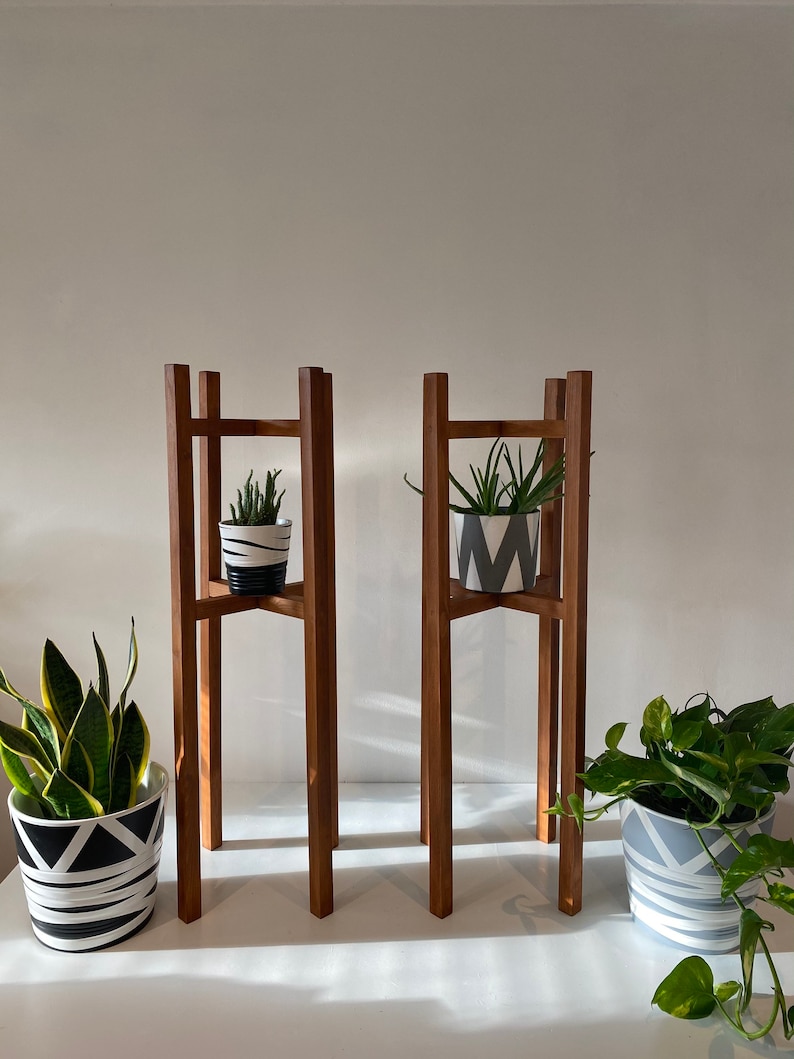 May include: Two wooden plant stands with three tiers each. The stands are made of dark brown wood and have a simple, modern design. Each stand has a potted plant on the top tier. The plants are in white pots with black geometric patterns.