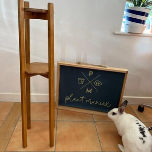 May include: A wooden plant stand with two shelves. The stand is made of light brown wood and has a simple design. The stand is in front of a black sign with the text "plant maniacs" in gold lettering.