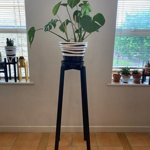 Puede incluir: Un soporte de planta negro con cuatro patas sostiene una planta en maceta con grandes hojas verdes. La maceta es blanca con rayas negras.