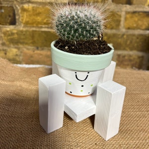 Pode incluir: Uma pequena planta de cacto em um vaso branco com uma borda verde e bolinhas pretas e verdes. O vaso tem um rosto sorridente pintado à mão. O vaso está sentado em um suporte de madeira branco com quatro pernas.