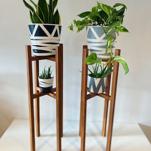 May include: Two wooden plant stands with three tiers each. The top tier of each stand holds a potted plant. The bottom tier of each stand holds a smaller potted plant. The plants are in white pots with black and grey geometric patterns.