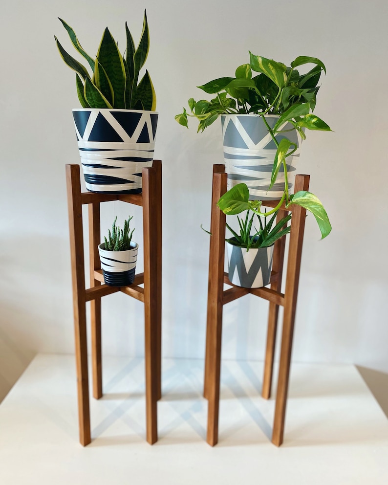 May include: Two wooden plant stands with three tiers each. The top tier of each stand holds a potted plant. The bottom tier of each stand holds a smaller potted plant. The plants are in white pots with black and grey geometric patterns.