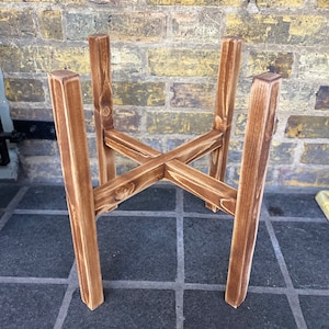 May include: A wooden plant stand with a simple, rustic design. The stand is made of four pieces of wood that are joined together in a cross shape. The stand is brown in colour and has a natural finish.