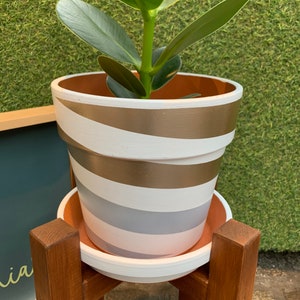 May include: A white planter with gold and gray stripes sits on a wooden plant stand. The planter has a terracotta rim and is filled with a green plant.