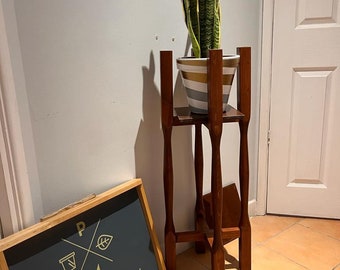 May include: A wooden plant stand with four legs and a shelf, supporting a potted snake plant with green and yellow leaves. The plant pot is white with gold and black stripes. A wooden sign with the text "Plant Maniax" is on the floor.