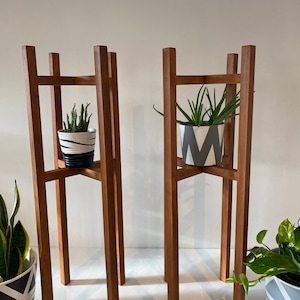 May include: Two wooden plant stands with a square base and four legs. Each stand has a shelf in the middle and is holding a potted plant.
