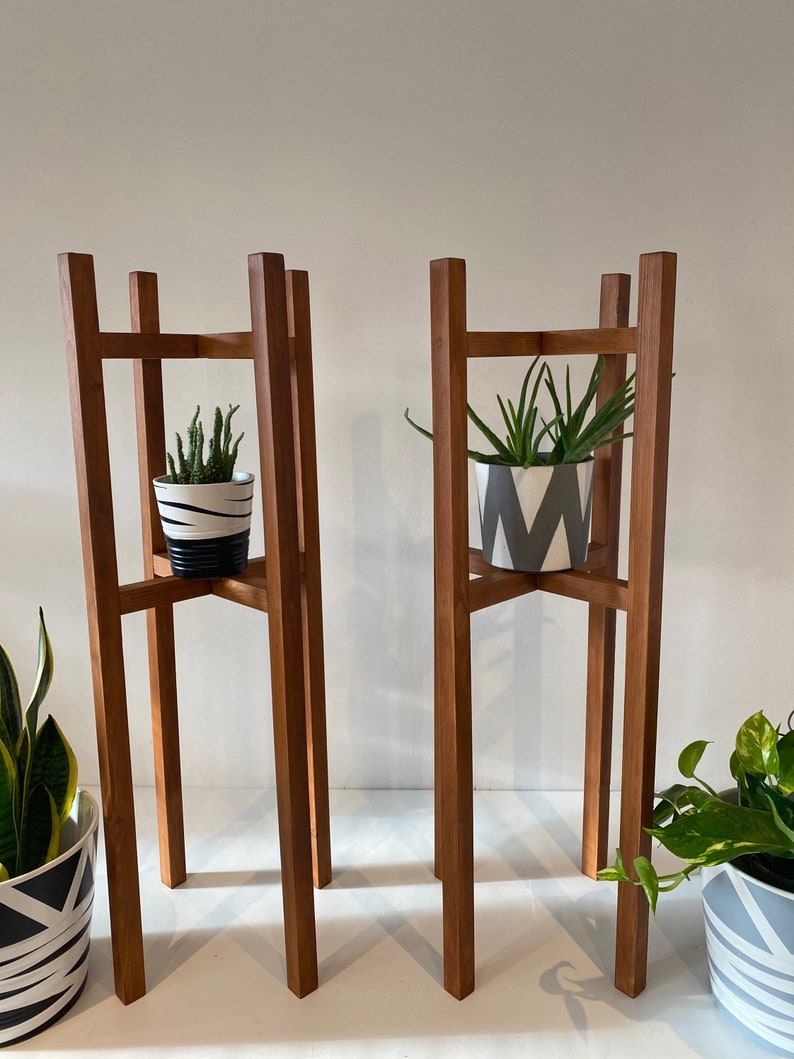 May include: Two wooden plant stands with a square base and four legs. Each stand has a shelf in the middle and is holding a potted plant.