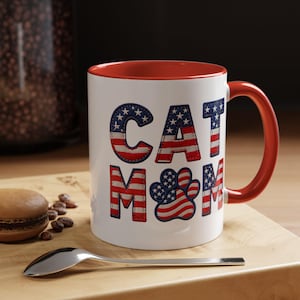 May include: A white ceramic mug with a red interior and handle. The mug features the text "CAT MOM" in a patriotic design with the American flag. A silver spoon and a pastry are on a wooden surface.