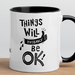 May include: White ceramic mug with a black interior and handle. The mug features the text "Things Will Probably Be OK!" in a black, handwritten-style font, along with a thumbs-up illustration. The mug is placed on a wooden surface.