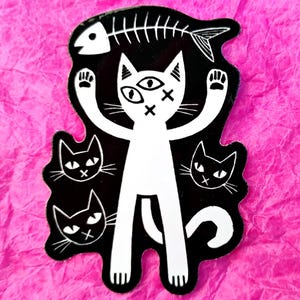 Op de afbeelding: Een zwart-witte sticker met een gestileerde kat met drie ogen en doorgestreepte ogen. De kat heft zijn poten op en heeft een vissenskelet erboven. Er zijn ook drie kleinere kattengezichten aanwezig.
