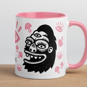 Puede incluir: Taza de cerámica blanca con borde y asa de color rosa. La taza presenta una ilustración en blanco y negro de una criatura de tres ojos con barba y una sonrisa de dientes. El fondo está decorado con setas y ojos rosas.