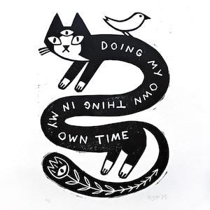 Op de afbeelding: Zwart-wit kunstprint met een gestileerde kat met drie ogen en een vogel. De kattenlichaam vormt een lus met de tekst "DOING MY OWN THING IN MY OWN TIME". Een snijgereedschap en houtsnippers zijn ook zichtbaar.