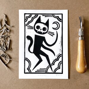 Può includere: Stampa a linoleum in bianco e nero di un gatto con testa di teschio, in piedi su nuvole con una luna crescente sullo sfondo. La stampa è intitolata "Mischief" e firmata "RSPP".