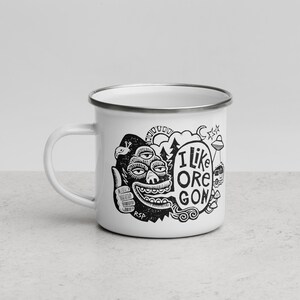 Puede incluir: Una taza de esmalte blanco con borde plateado que presenta un estampado gr&aacute;fico negro de una criatura de tres ojos haciendo un gesto de pulgar hacia arriba, un globo de texto con I LIKE OREGON, sol, &aacute;rboles, un OVNI, caravana y setas.