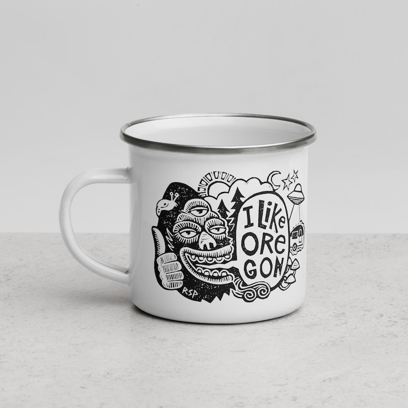 Puede incluir: Una taza de esmalte blanco con borde plateado que presenta un estampado gr&aacute;fico negro de una criatura de tres ojos haciendo un gesto de pulgar hacia arriba, un globo de texto con I LIKE OREGON, sol, &aacute;rboles, un OVNI, caravana y setas.
