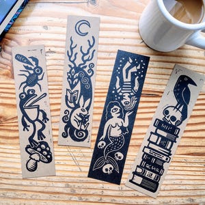May include: Set of four black and white illustrated bookmarks. The bookmarks feature a frog, a creature with antlers, a mermaid, and a crow perched on a stack of books. The bookmarks are printed on brown paper.