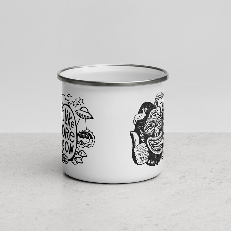 Puede incluir: Una taza de esmalte blanco con borde plateado que presenta ilustraciones gr&aacute;ficas en negro. El dise&ntilde;o incluye una criatura de tres ojos haciendo un gesto de pulgar hacia arriba, un OVNI, una caravana y el texto I LIKE OREGON en una fuente circular.