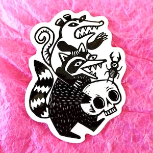 May include: Black and white sticker featuring a raccoon, opossum, and beetle with a skull. The raccoon holds the skull, while the opossum and beetle are positioned above. The sticker is on a pink, textured background.