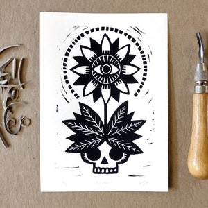 May include: Black and white linocut print featuring a stylized flower with an eye at the center, atop a stem with leaves and a skull. A carving tool and metal shavings are next to the print. The print is on a white paper.
