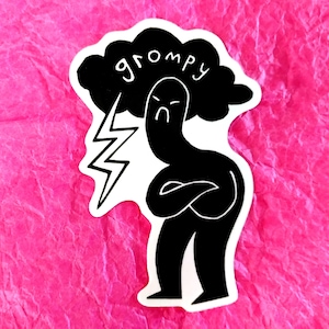 Może przedstawiać: Czarno-biała naklejka przedstawiająca zrzędliwą postać z kreskówki ze skrzyżowanymi ramionami, pod chmurą z napisem "grompy" i błyskawicą. Naklejka znajduje się na różowym tle.