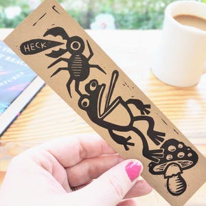 Puede incluir: Un marcapáginas marrón con un diseño de linograbado negro que presenta un insecto con la palabra «HECK» en un bocadillo, una rana y una seta. El marcapáginas se sostiene en una mano, con un libro y una taza de café en el fondo.