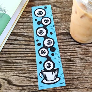 May include: A blue bookmark with a black and white coffee-themed design. The design features a series of stylized coffee cups and floral elements. The bookmark is next to a book and a glass of iced coffee.