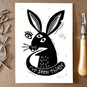 以下が含まれることがあります： 3つの目をしたウサギの白黒の版画。ウサギは直接鑑賞者を見ており、大きく歯の見える笑顔をしています。「I've seen things」というテキストがウサギの下に書かれています。右下にアーティストの署名「RSP'23」があります。