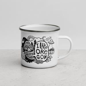 Puede incluir: Una taza de esmalte blanco con borde plateado y un estampado gr&aacute;fico negro. El dise&ntilde;o presenta una criatura de tres ojos haciendo un gesto de pulgar hacia arriba, un globo de texto con I LIKE OREGON, un sol, una luna, estrellas, un ovni y setas.