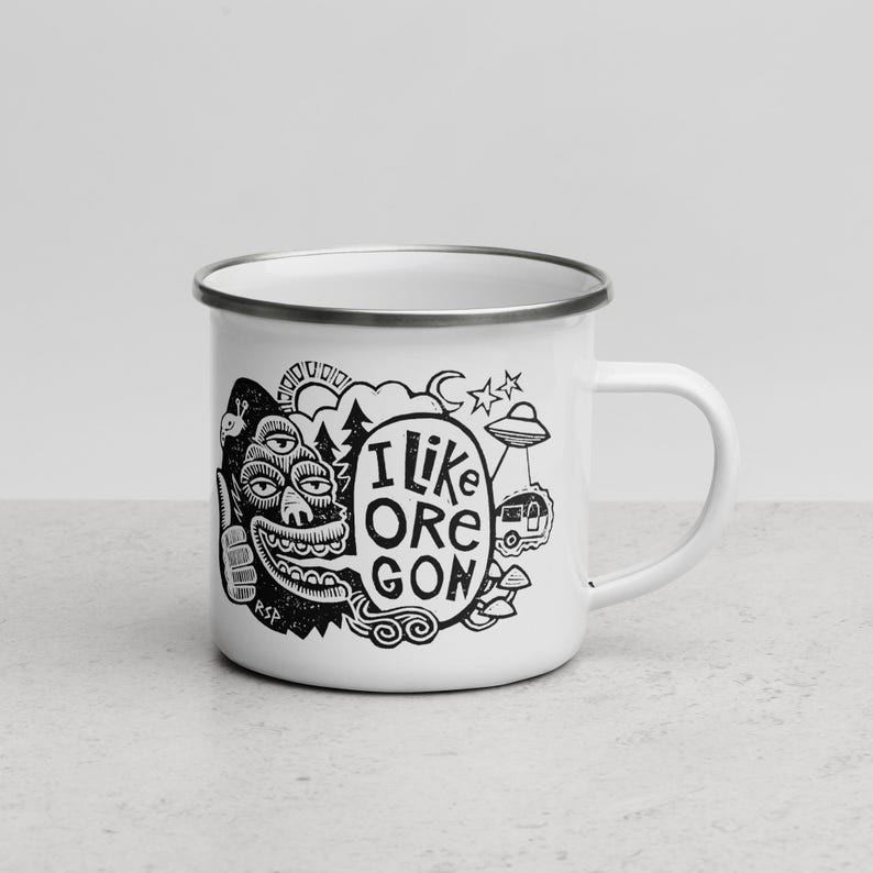 Puede incluir: Una taza de esmalte blanco con borde plateado y un estampado gr&aacute;fico negro. El dise&ntilde;o presenta una criatura de tres ojos haciendo un gesto de pulgar hacia arriba, un globo de texto con I LIKE OREGON, un sol, una luna, estrellas, un ovni y setas.