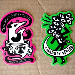 May include: Two die-cut stickers. One is pink and black with the text "TOO MUCH EVERYTHING ALL THE TIME" and a badger-like creature. The other is green and black with the text "I MADE IT WEIRD" and a figure holding a bouquet of eyes.