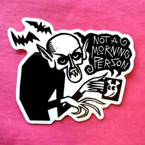 May include: A black and white sticker depicting a vampire holding a picture with a smiling face. The sticker has the text "NOT A MORNING PERSON" in a speech bubble, with bats in the background. The sticker is on a pink background.
