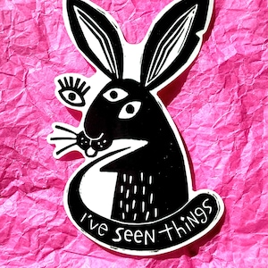 May include: Black and white illustration of a rabbit with three eyes and a tongue sticking out. The rabbit is wearing a white collar with the text "I've seen things".